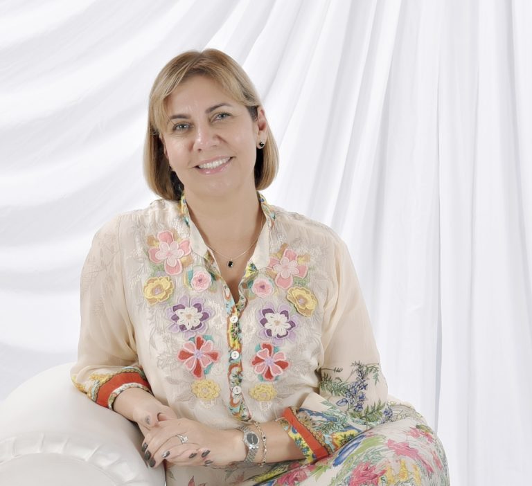 A woman wearing floral attire and jewelry, seated against a white background, striking a pose